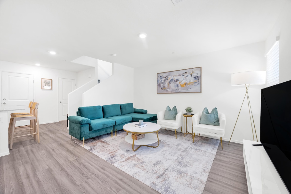 A modern living area features a teal sectional sofa accompanied by two white armchairs, creating a comfortable seating arrangement. A round coffee table sits at the center, and a large area rug adds texture to the space. Natural light brightens the room through nearby windows.