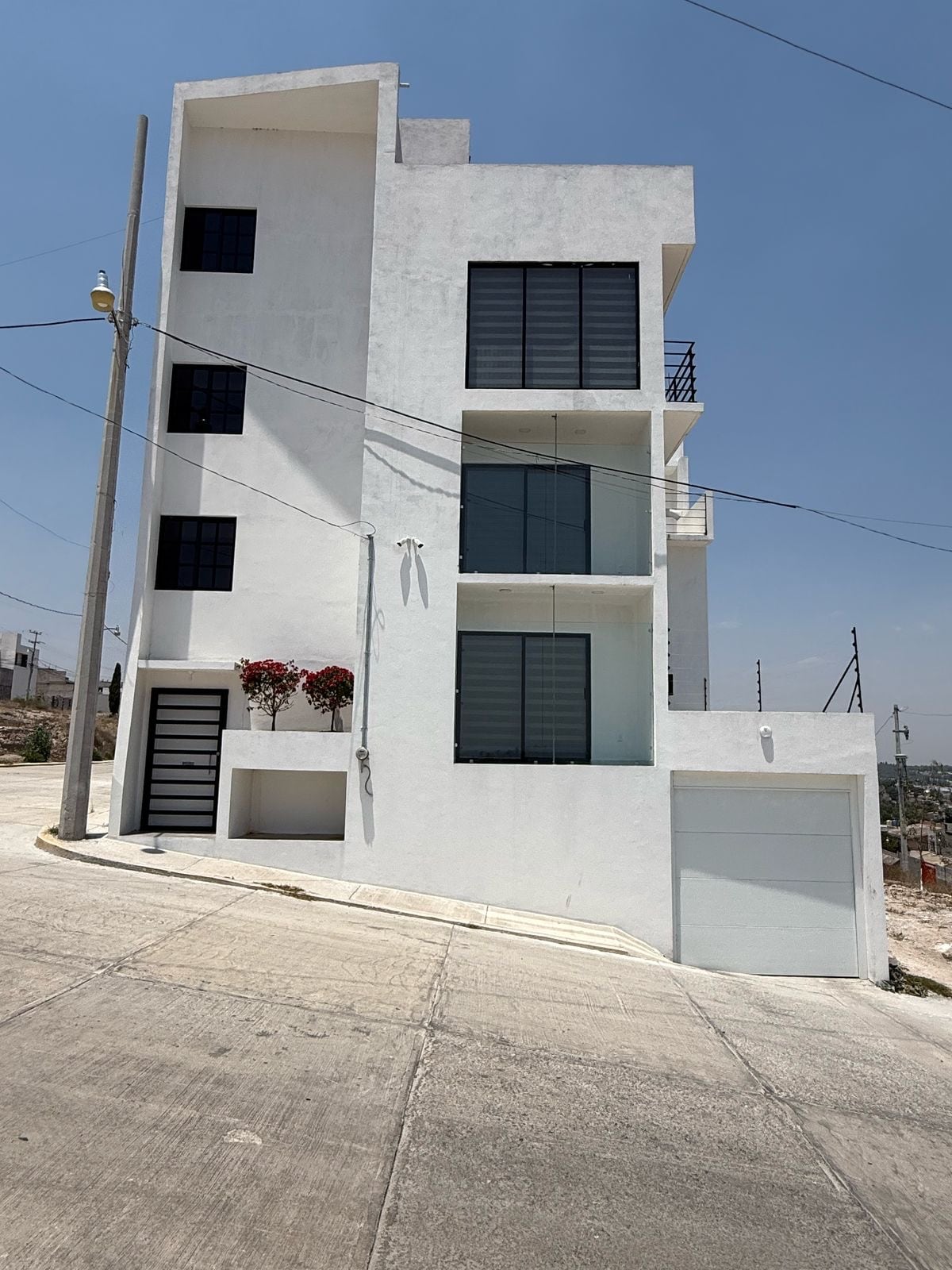 A modern three-story building is displayed, featuring a clean white exterior with large windows. A garage door is visible at the base, while flower pots add a touch of color to the façade. The surrounding area is characterized by a sloped street.