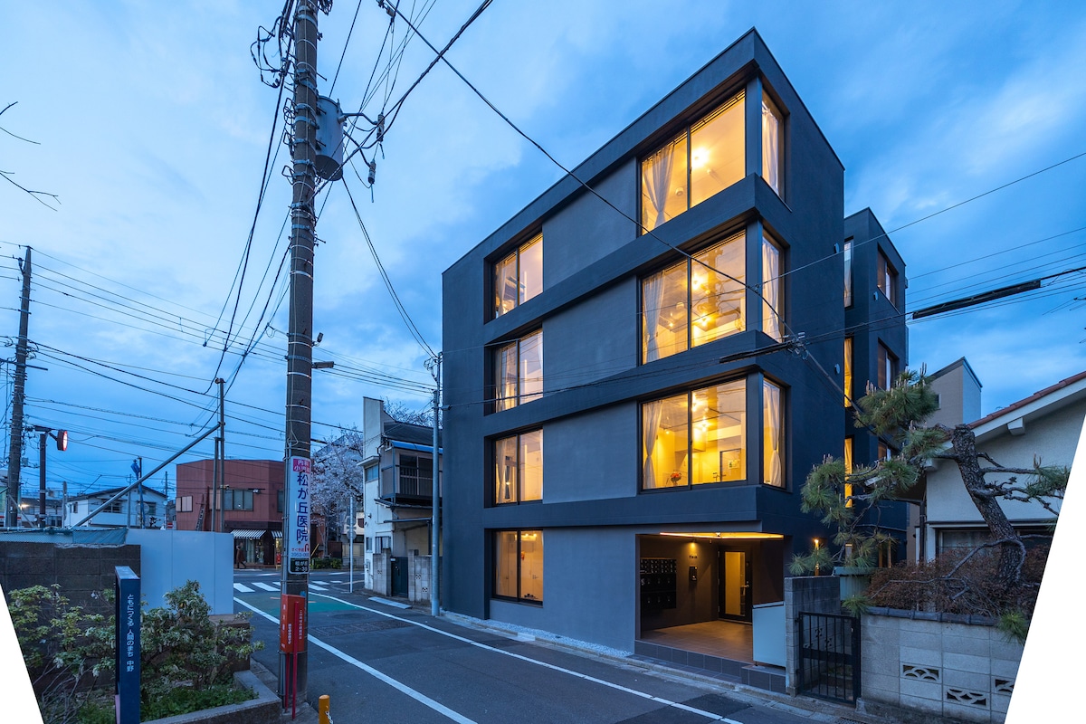 A modern four-story building features large windows that allow light to illuminate the interior spaces. Street elements, including power lines and sidewalk, can be seen alongside the smooth exterior of the structure. The surrounding area reflects a blend of urban and residential characteristics.