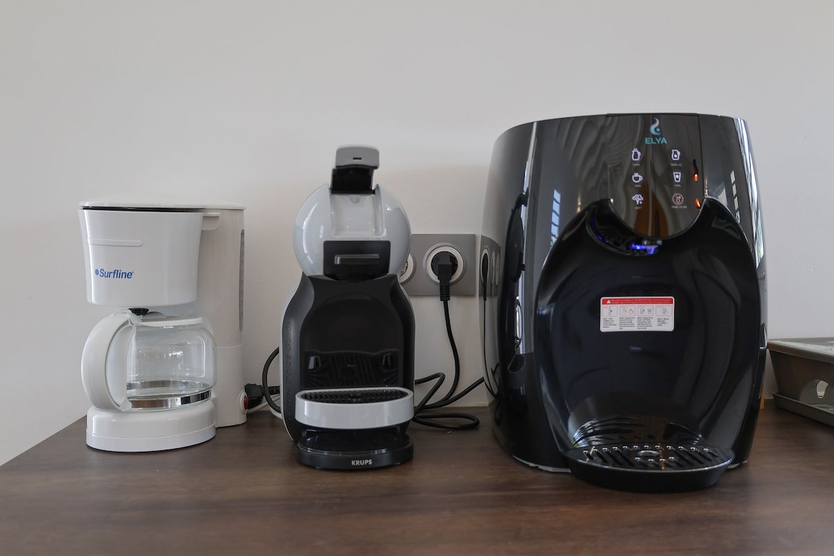 A countertop displays three coffee-making appliances: a white traditional coffee maker, a sleek grey pod coffee machine, and a modern black water dispenser. The devices are plugged in and organized neatly, with electrical outlets visible in the background.