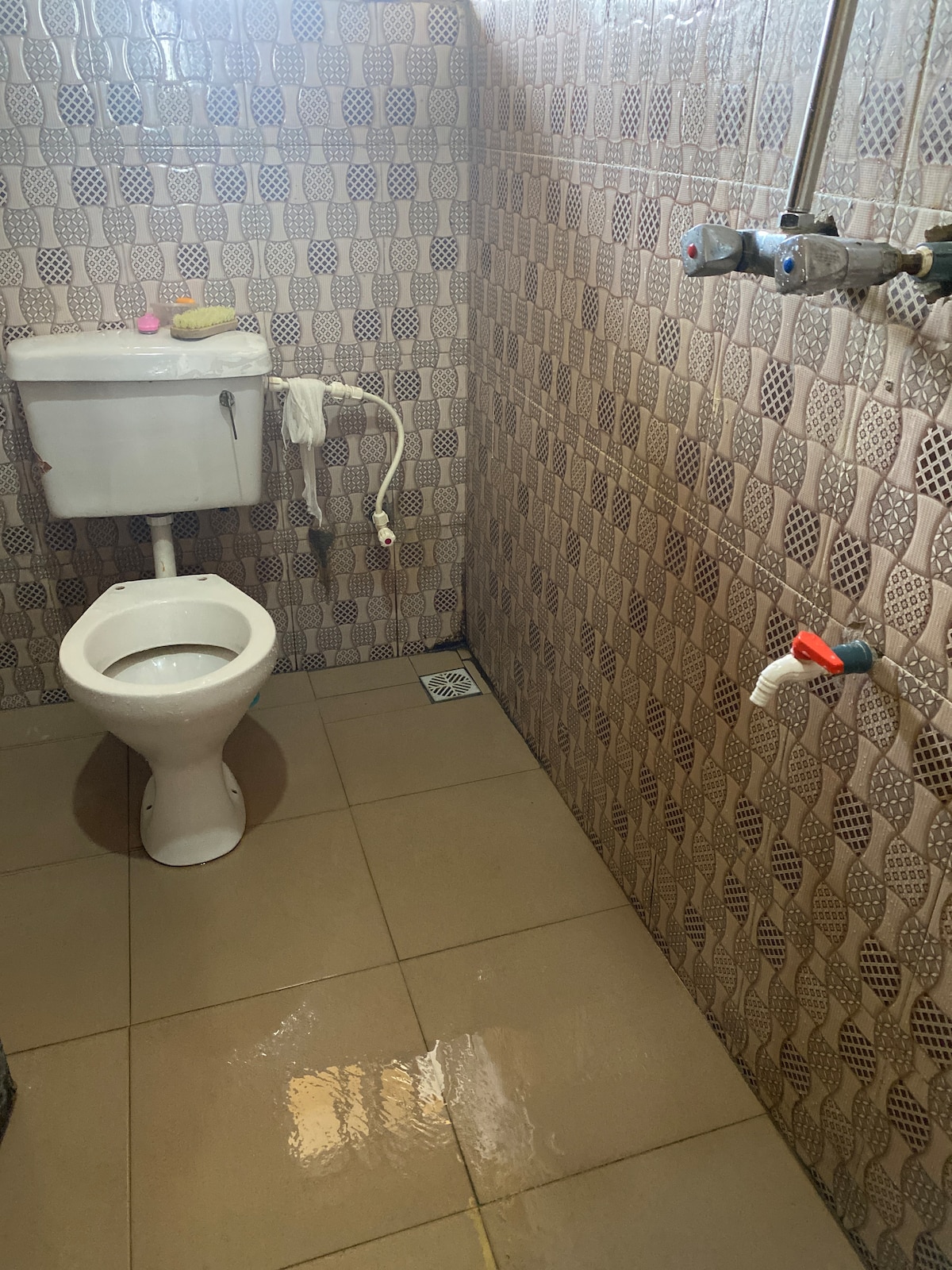 The bathroom features a standard toilet beside a wall-mounted sink. The tiled walls display a subtle pattern, and the floor is finished with large tiles. A faucet is visible on the right, indicating a functional space designed for convenience.