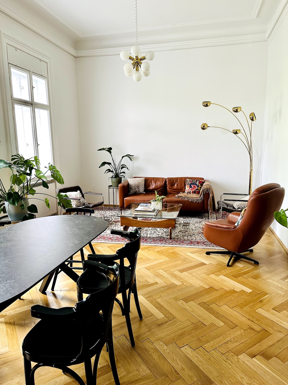 The open plan lounge area features a rich wooden floor with a geometric pattern. A brown leather couch sits against the wall, accompanied by a lounge chair and sleek floor lamp. A dining table with black chairs occupies the space, accented by vibrant plants and decorative elements.