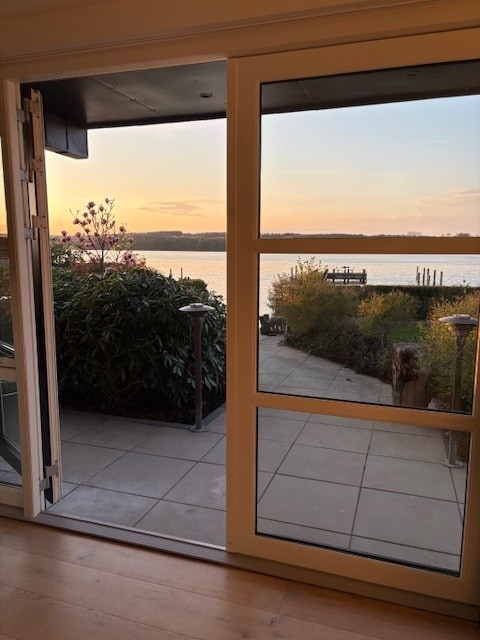 A spacious doorway opens to a patio area, featuring lush greenery and outdoor lighting. The view beyond includes a serene body of water reflecting the soft hues of a sunset. A pathway leads to a dock, creating a peaceful connection to nature.