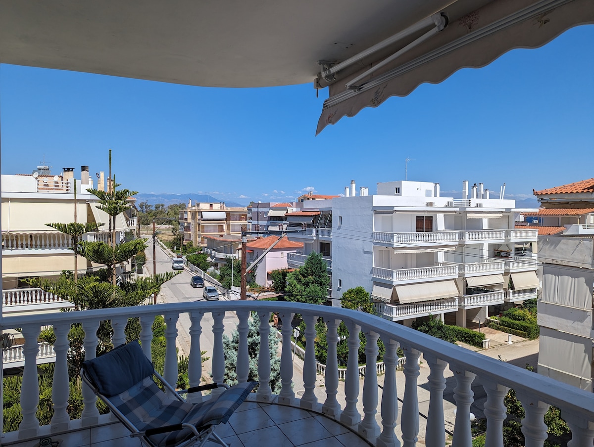 A spacious balcony is visible, featuring a comfortable chair and railing with decorative balusters. The scene includes neighboring buildings, green landscaping, and a clear blue sky. Distant mountains are subtly seen in the background, completing the serene view.