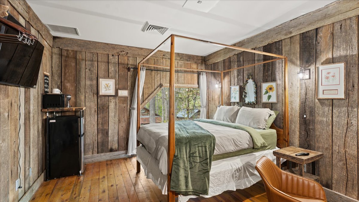 A cabin suite is characterized by rustic wood-paneled walls and warm lighting. A four-poster queen bed with soft linens is positioned centrally. A mini fridge and coffee maker are visible on one side, while a seating area is set up with a chair and small table nearby.