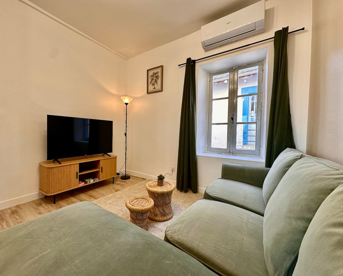 A cozy living area features a large green sofa and a wooden TV stand with a flat-screen television. Two small round tables made of natural materials are placed side by side. Light flows in through a window adorned with dark green curtains, enhancing the inviting ambiance.