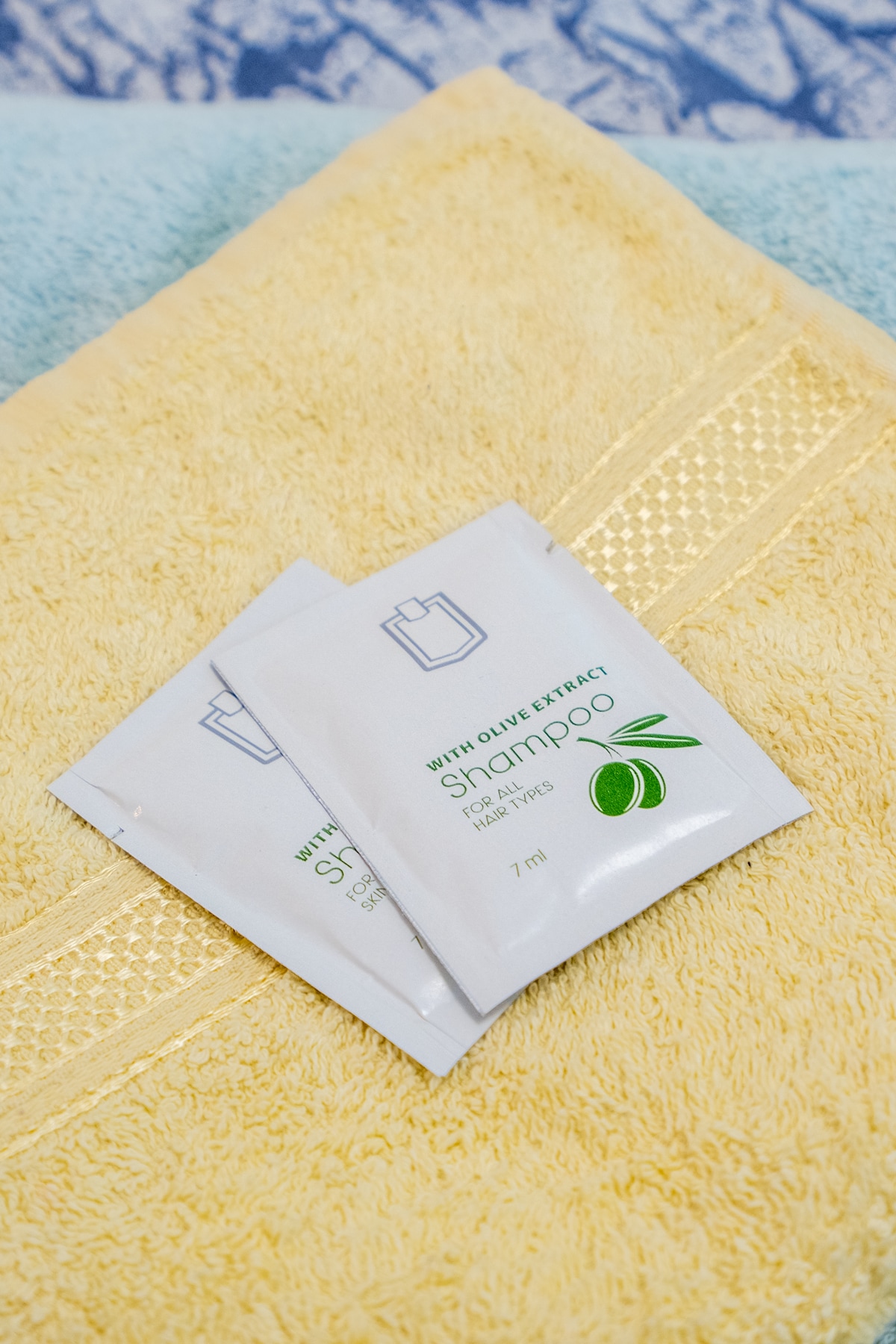 Two packets of shampoo with olive extract are placed on a textured yellow towel. The vibrant colors of the towels create a welcoming atmosphere, enhancing the bathroom essentials provided for guests.