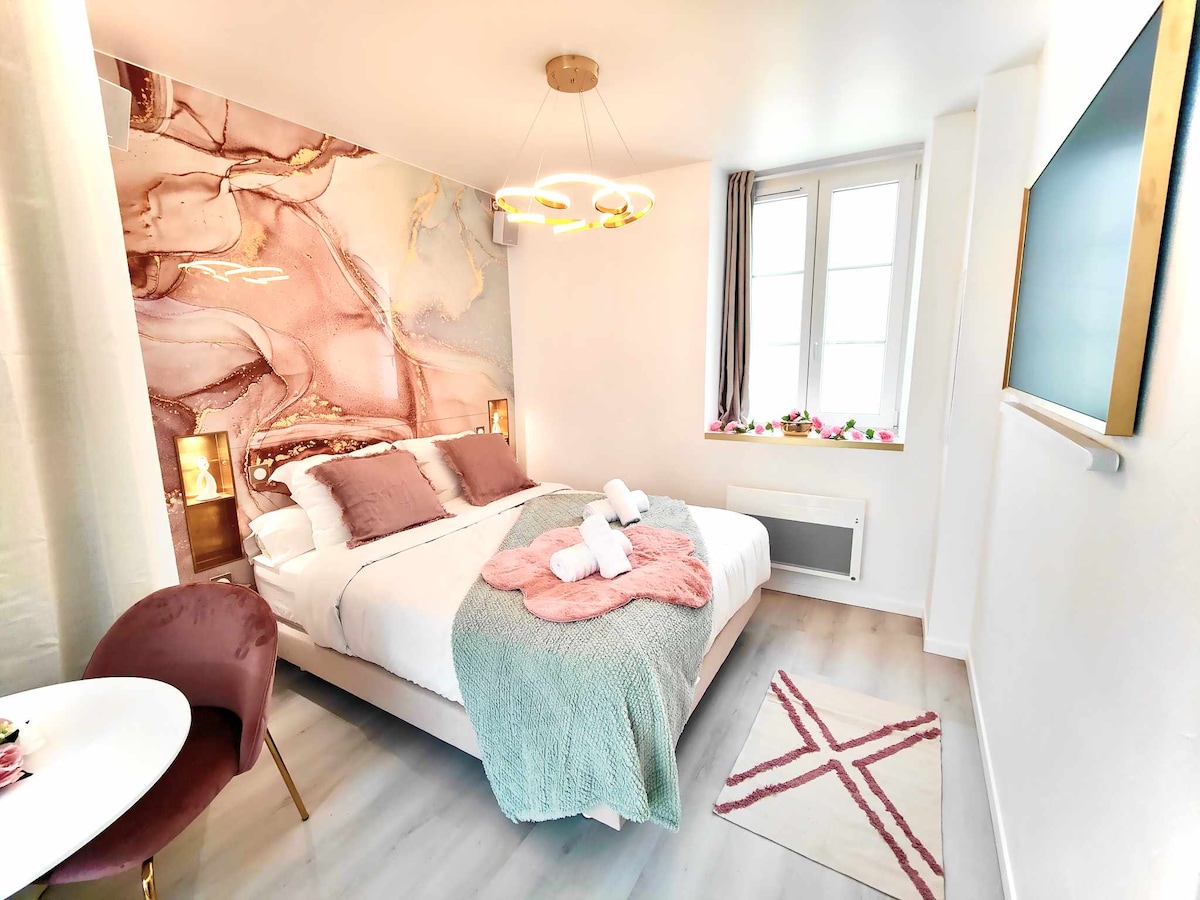 A cozy studio bedroom features a stylish bed adorned with soft pillows and a textured throw. Elegant lighting fixtures illuminate the space, while a delicate wall mural adds visual interest. A small dining table and chair are positioned nearby, with a colorful area rug underfoot.