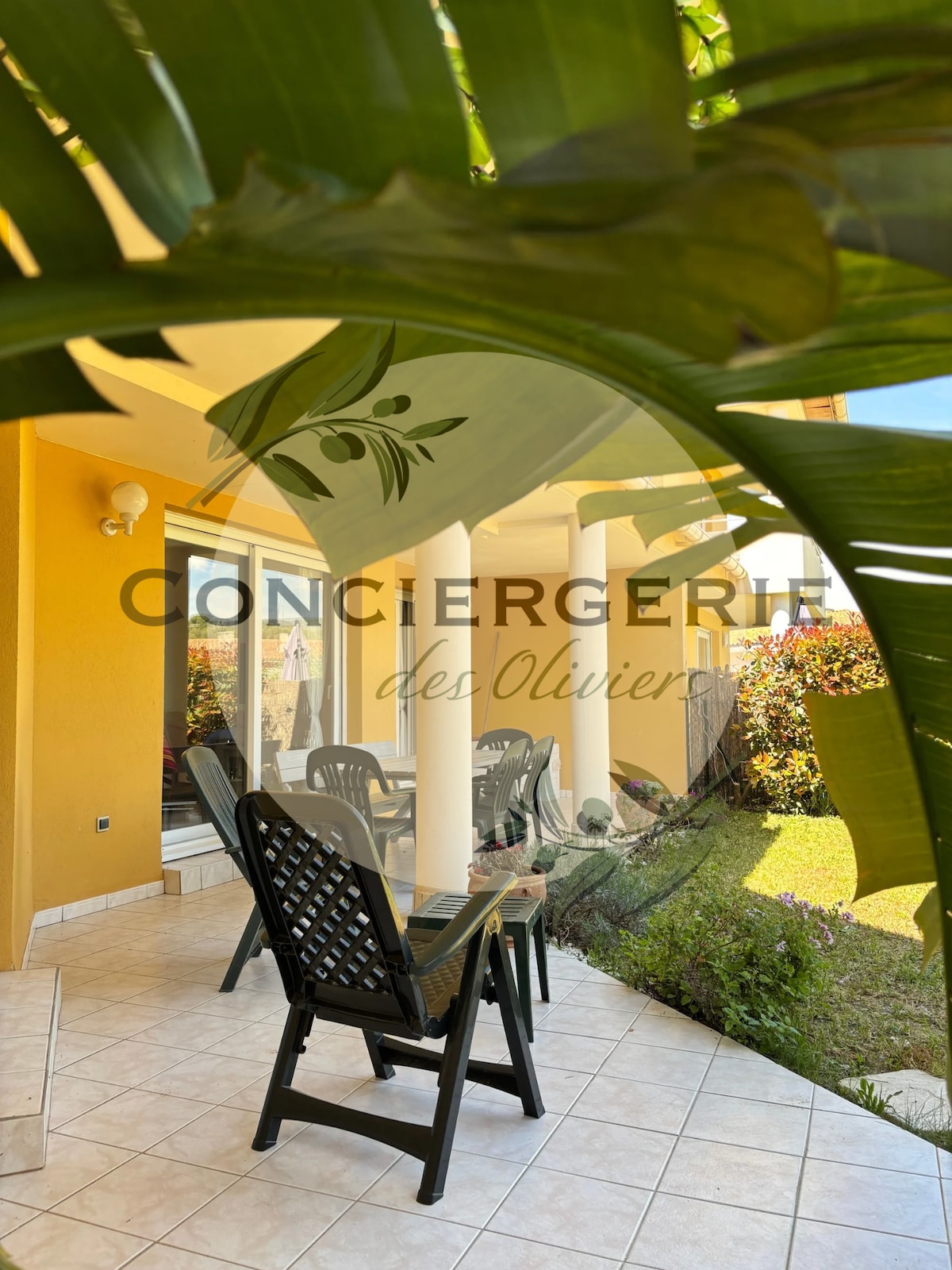 A shaded patio area is seen, featuring four dark outdoor chairs arranged around a small table. The space is framed by tall green plants, providing a sense of privacy, while bright flowers and well-maintained grass are visible in the background.