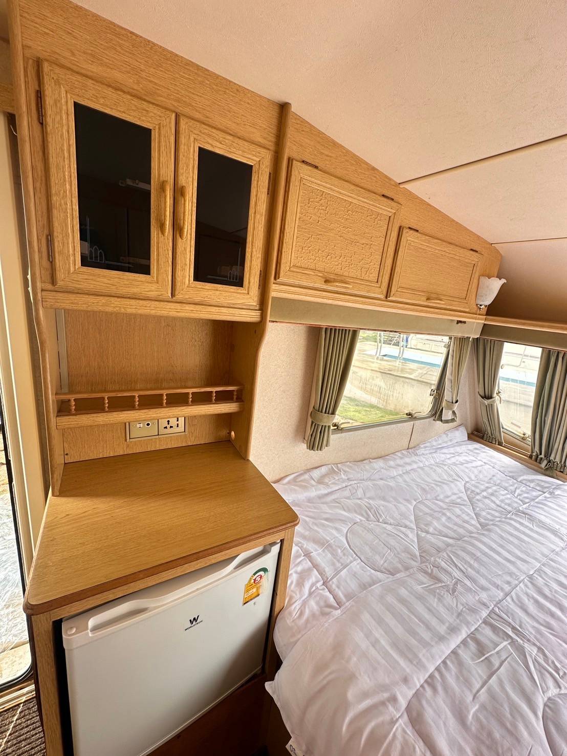 A cozy interior of a caravan features a wooden storage cabinet above a compact refrigerator. A spacious bed with crisp white bedding is visible, accompanied by large windows allowing natural light to fill the room. Soft curtains add a touch of comfort to the atmosphere.