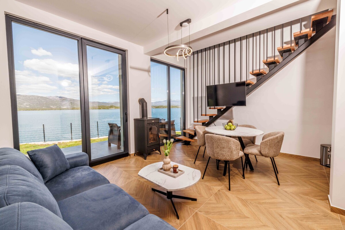 A modern living area features a comfortable blue sofa facing a stylish round table surrounded by four upholstered chairs. Large glass windows offer a scenic view of the lake, while a staircase with wooden steps and a sleek railing leads to the upper level.