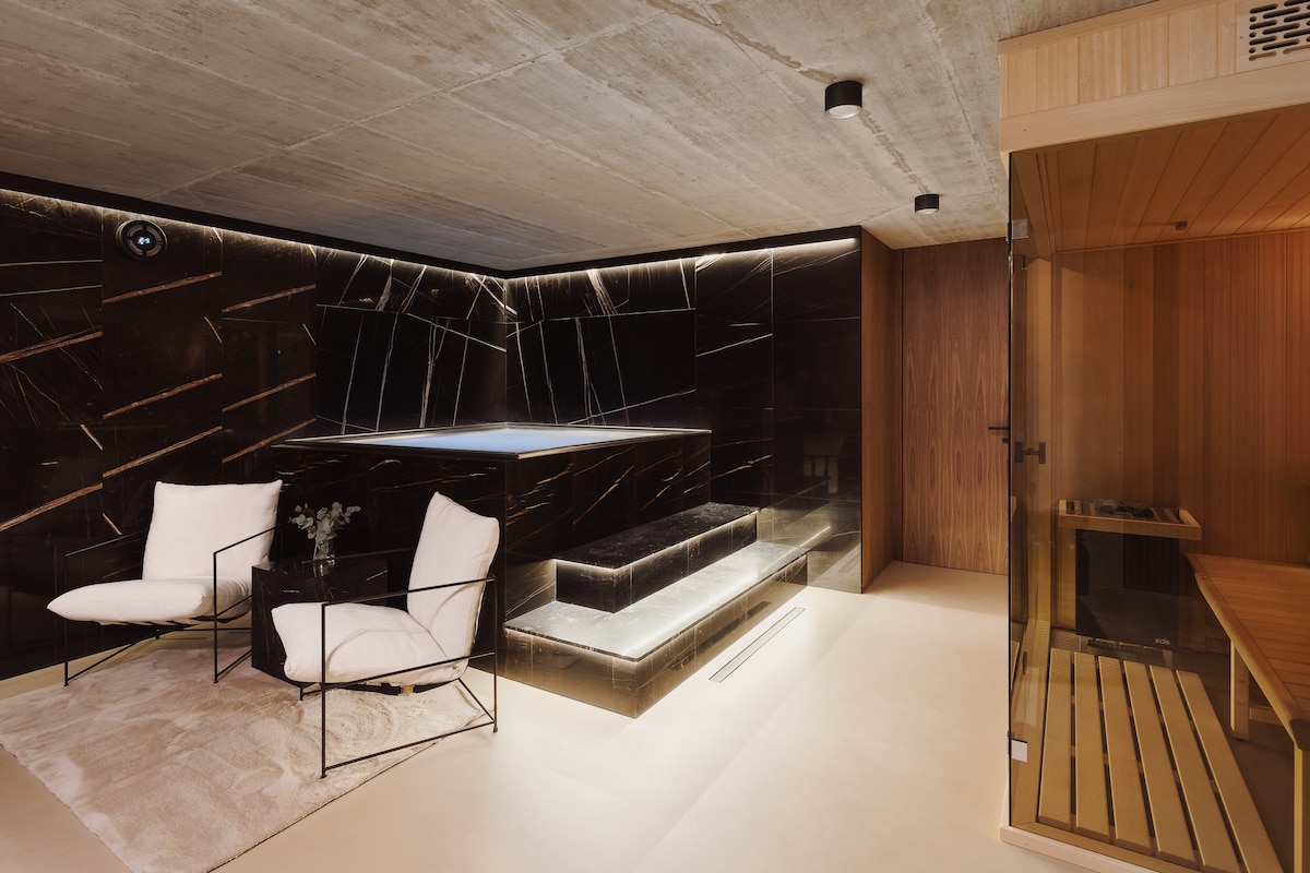A modern wellness area features a sleek black tile design with subtle lighting. A small relaxation pool is complemented by two white armchairs on a soft rug. A wooden sauna is positioned in the corner, enhancing the calming atmosphere of the space.