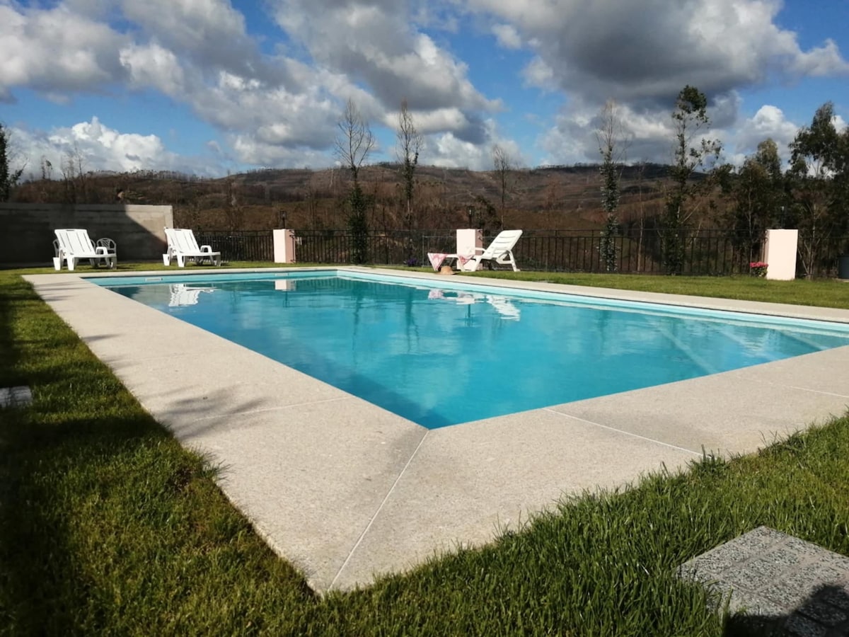 A rectangular swimming pool is surrounded by well-maintained grass, with two white lounge chairs positioned on one side. The pool reflects the blue sky and clouds, while lush trees and gentle hills are visible in the background.