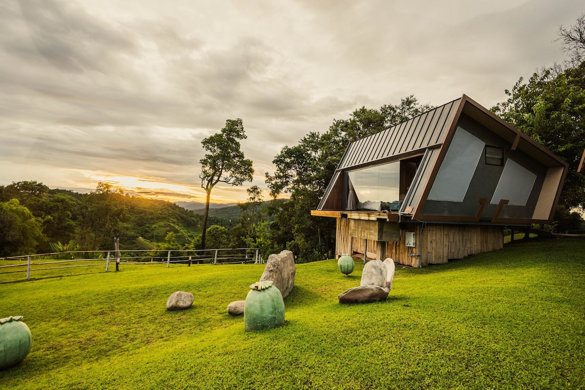 A modern retreat is positioned on a grassy landscape, featuring a sloped metal roof and large windows that allow for a panoramic view of the surrounding hills and sunset. Natural stone formations are scattered across the lawn, enhancing the connection to the serene environment.