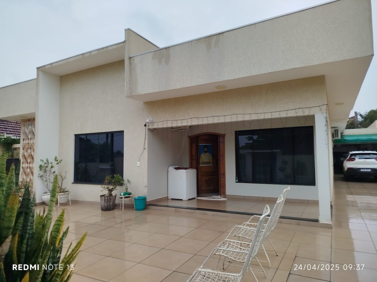 A modern exterior of a single-story residence is presented, featuring large windows that allow natural light to illuminate the interior. A covered entrance leads to the front door, while potted plants and white chairs add a touch of greenery to the paved patio area.