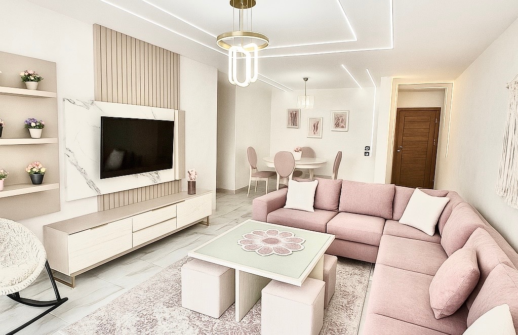 A comfortable living area features a large pink sectional sofa arranged around a coffee table with a floral design. A minimalist entertainment unit supports a television, while soft lighting from the ceiling highlights the pastel decor. A dining area with round, cushioned chairs is visible in the background.