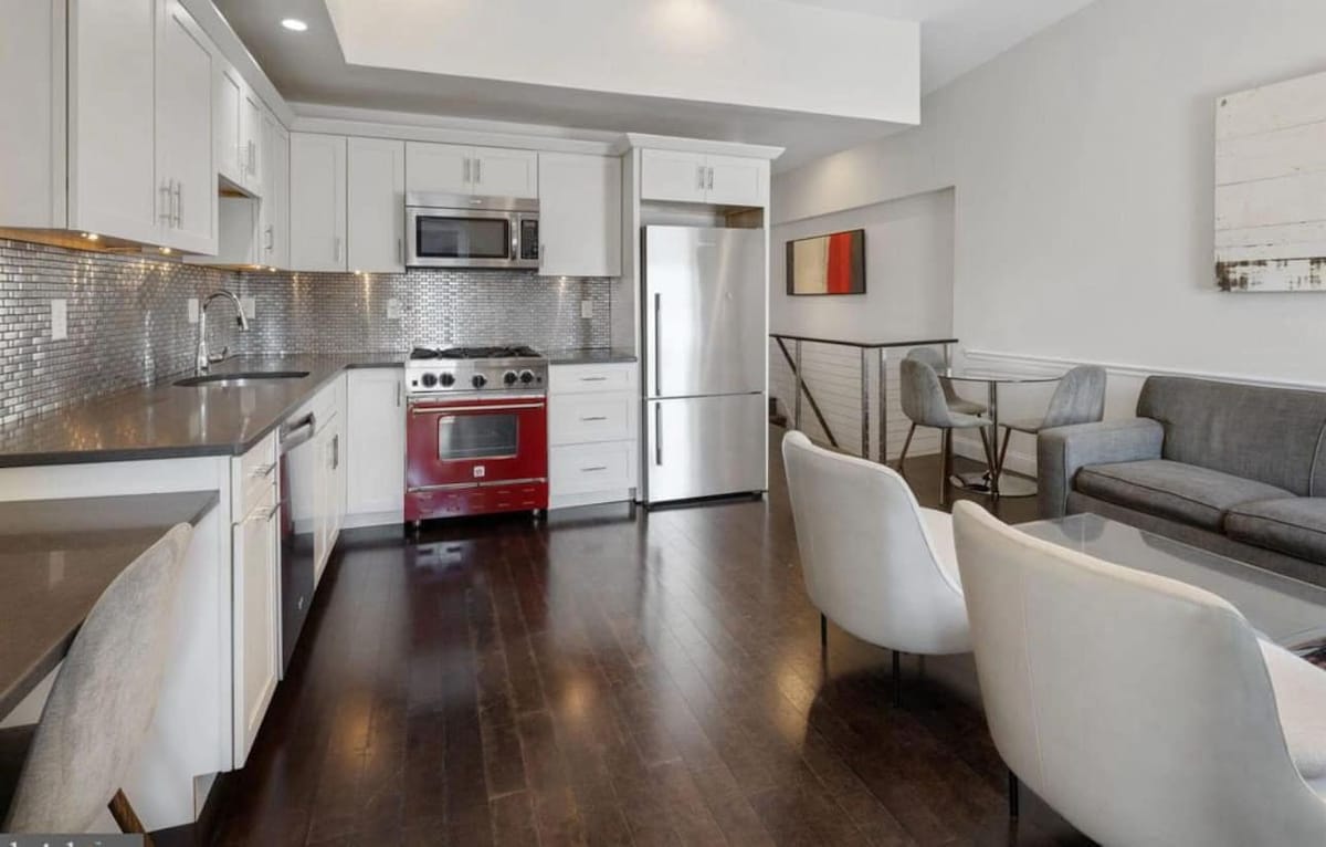 A modern kitchen and dining area feature sleek cabinetry and stainless steel appliances. A red gas stove adds a vibrant touch against the neutral tones. A comfortable seating area with a gray sofa is positioned near a staircase, with elegant dining chairs arranged around a round table.