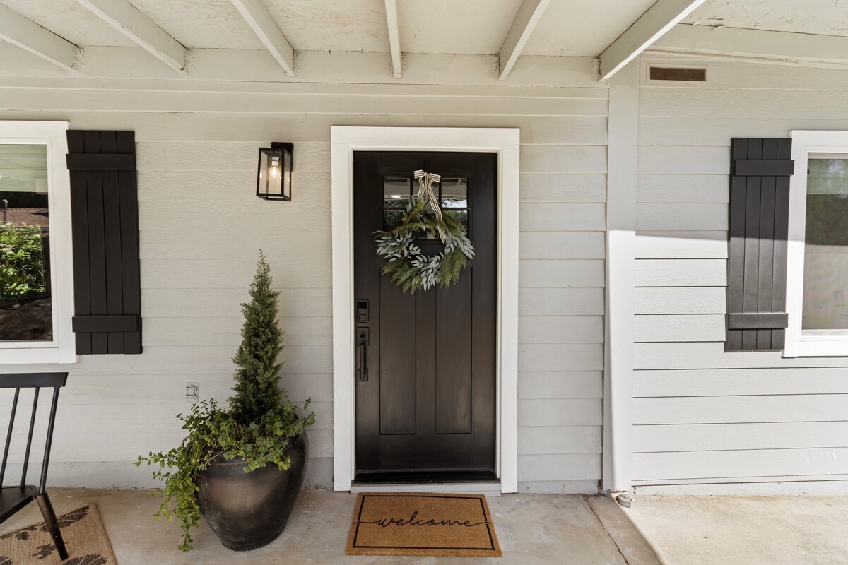 The entrance features a modern black door adorned with a decorative wreath. A welcoming doormat reads 'welcome.' A neatly trimmed evergreen plant is placed in an elegant pot, accompanied by a small black table against the house's exterior, showcasing a pleasant and inviting entryway.
