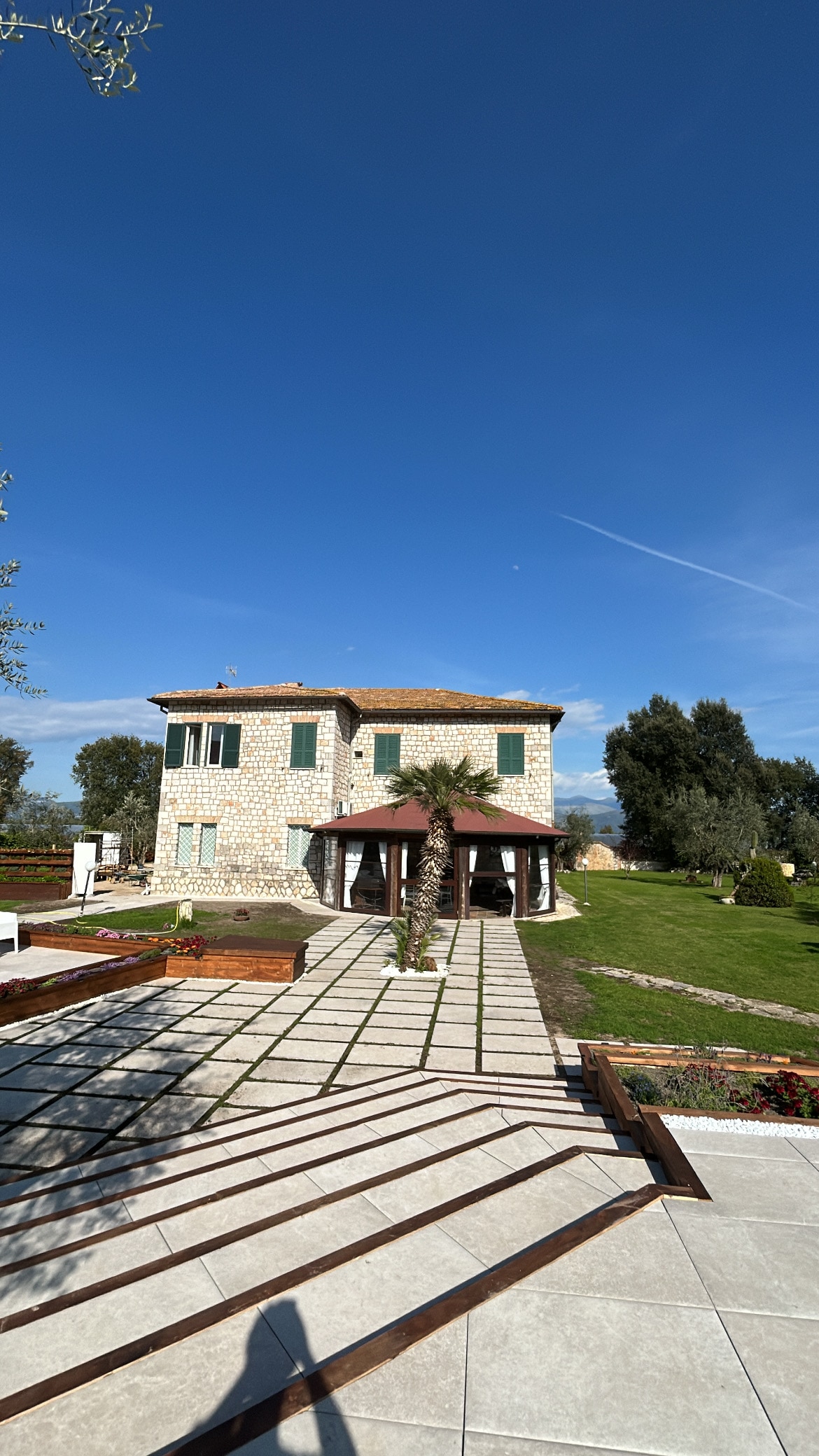 A two-story stone house is seen, featuring multiple green shuttered windows and a tiled roof. A pathway lined with stone tiles leads towards the entrance, flanked by landscaped greenery and a palm tree, under a clear blue sky.