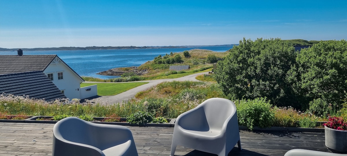 A spacious terrace features modern gray chairs, offering a view of the serene water and lush landscape. Clear skies enhance the scenery, while the inviting space is surrounded by greenery and vibrant flowers.