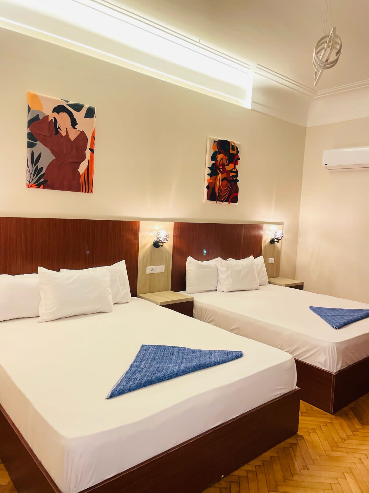 A spacious sleeping area is furnished with two beds, each arranged with white linens and a blue accent blanket. Artwork hangs on the light-colored walls, and bedside lighting is positioned for convenience. An air-conditioning unit is mounted above, ensuring a comfortable environment.