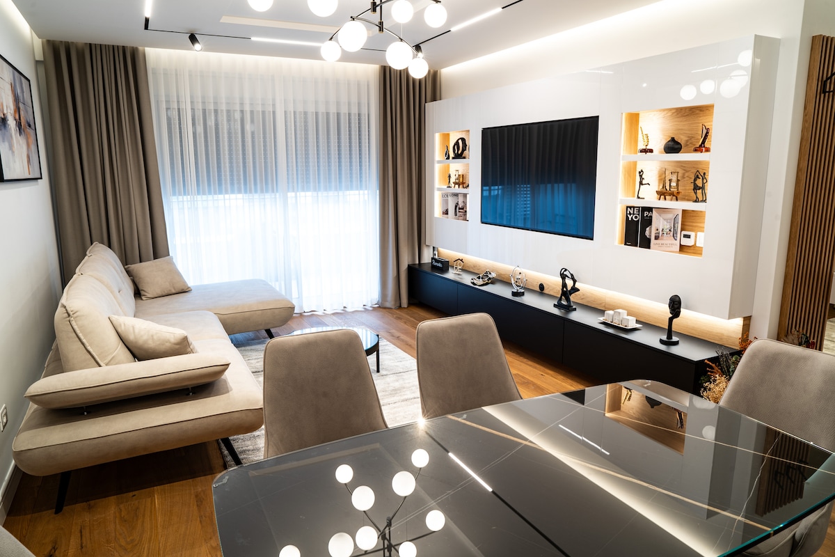 A modern living area features a spacious beige sectional sofa positioned beside a large window, providing natural light. A wall-mounted television is centered on a sleek black media unit, which displays decorative items. The dining area is visible with an elegant glass table and stylish chairs.