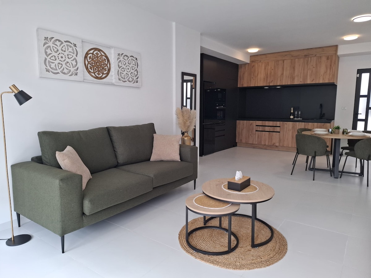 A modern living space is showcased with a green sofa and a set of circular coffee tables placed on a textured rug. The kitchen area features dark cabinetry against light walls, while a dining table with stylish chairs is visible in the background.