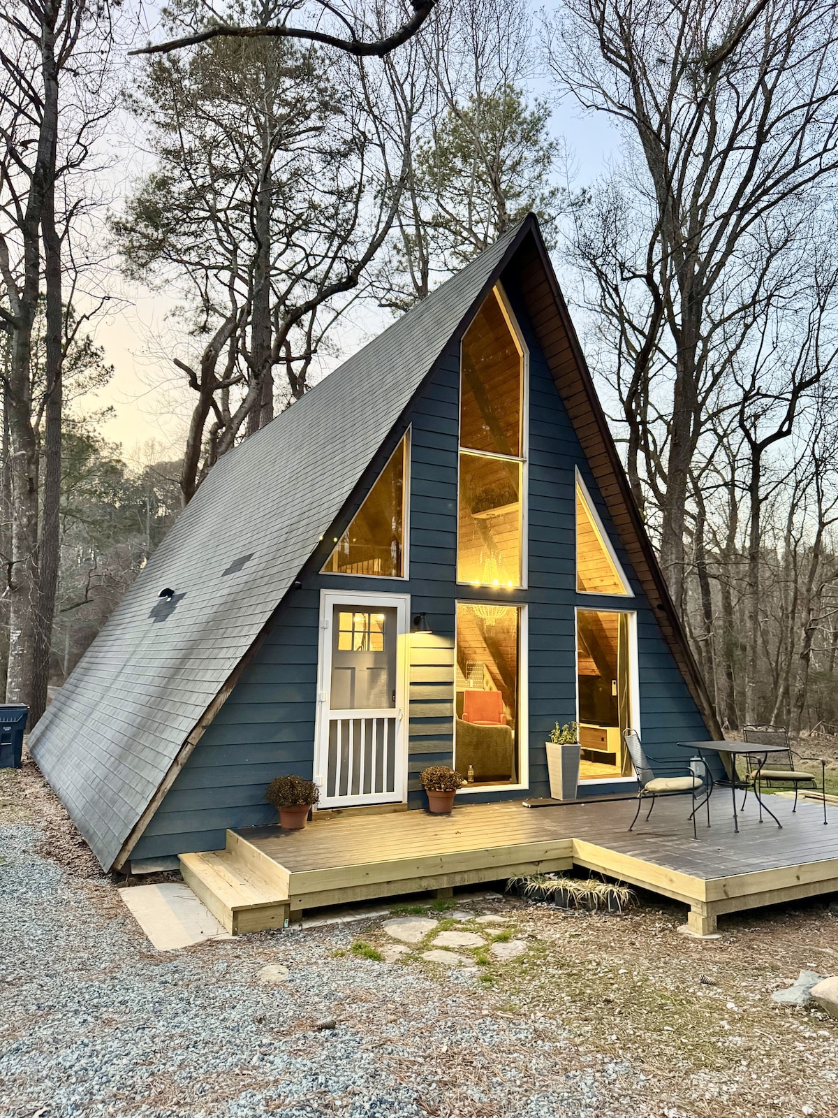 An A-Frame cabin showcases a striking triangular design with expansive glass windows reflecting the warm glow of interior lighting. A wooden deck leads to an entrance adorned with potted plants, with gravel paths and trees providing a serene natural backdrop.