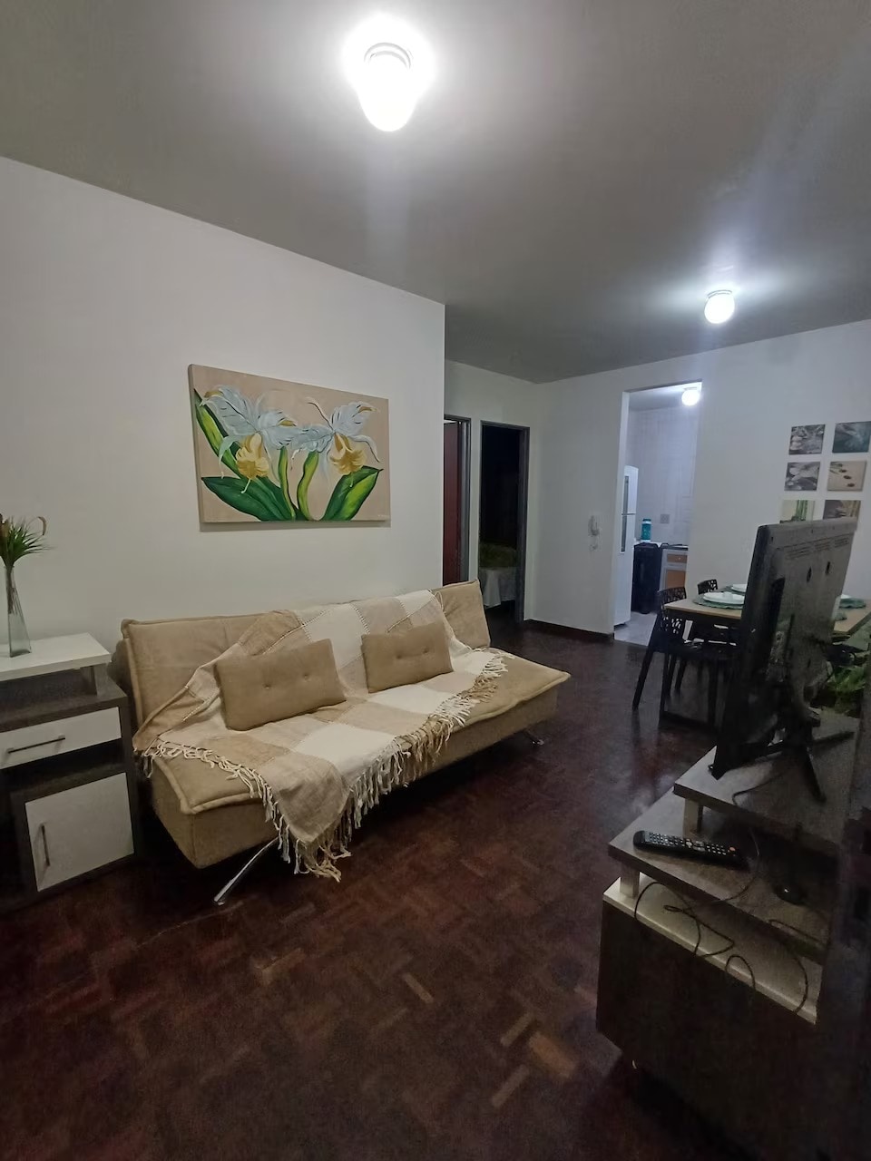 A cozy living space is presented, featuring a light-colored sofa adorned with neutral throw pillows and a woven blanket. A piece of wall art depicting flowers is displayed above the sofa. A dining area is visible in the background, along with a television unit.