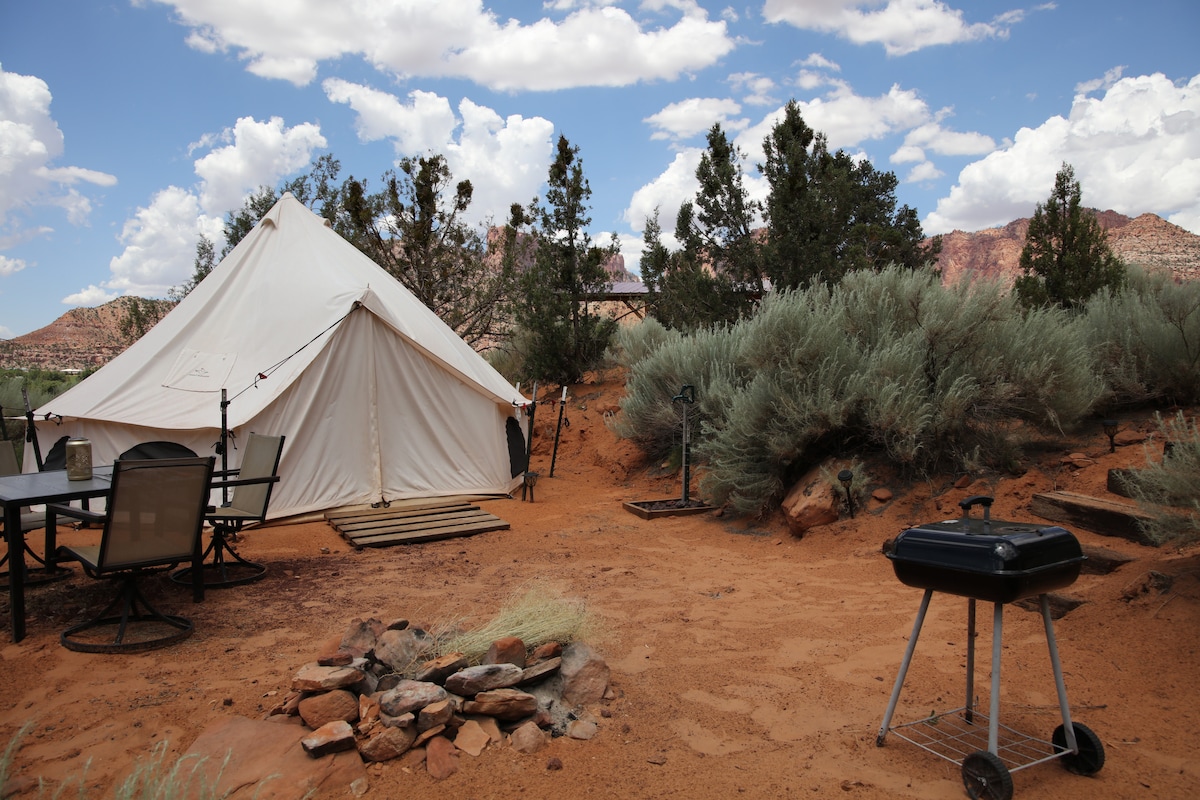 A spacious canvas tent is situated on a sandy area, surrounded by desert vegetation and distant rock formations. A small table with chairs is positioned nearby, and a BBQ grill rests on the ground, adjacent to a fire pit crafted from stones.