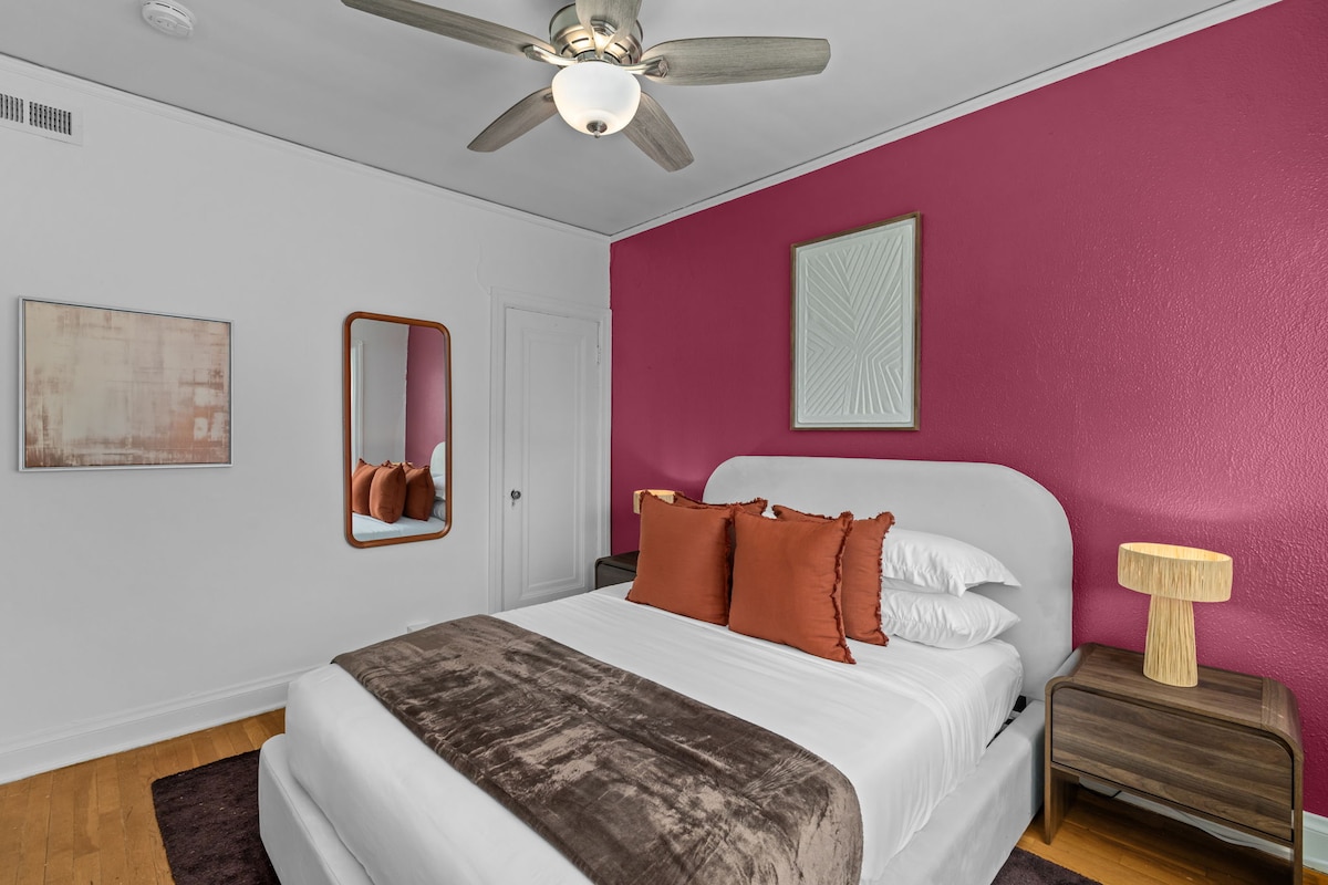 A well-appointed bedroom features a vibrant pink accent wall, complemented by a neutral bed with plush bedding and decorative orange pillows. A stylish wooden nightstand holds a unique lamp, while a mirror adds depth to the space. Natural light enhances the cozy atmosphere.