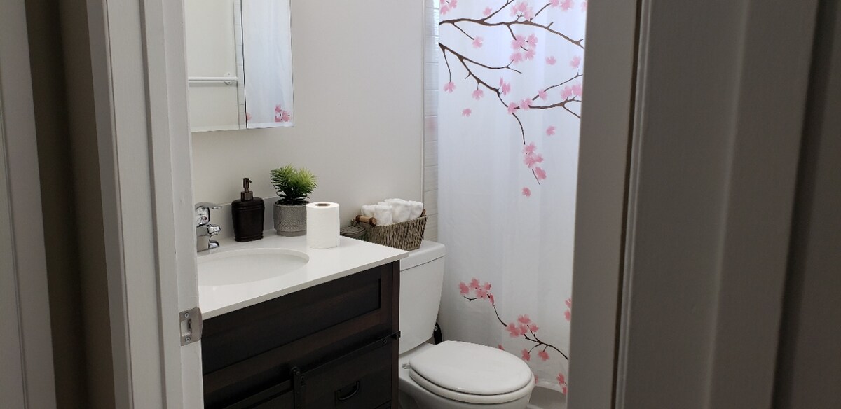 A well-appointed bathroom features a clean vanity with a mirror above and ample counter space. A soft floral shower curtain adorns the tub, complemented by potted plants and neatly organized towels. Natural light is allowed in through the window, enhancing the serene environment.