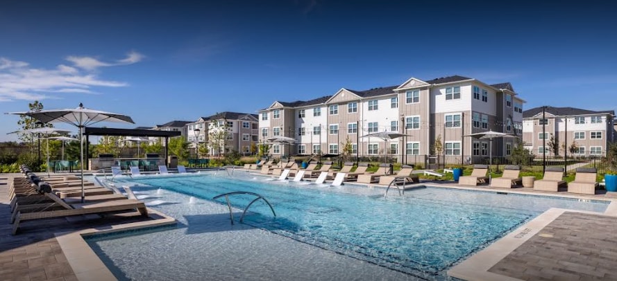 A modern outdoor pool area features a clear blue swimming pool surrounded by lounge chairs and umbrellas. The well-maintained landscape includes green foliage and seating areas, while contemporary apartment buildings provide a backdrop, creating an inviting space for relaxation and leisure.