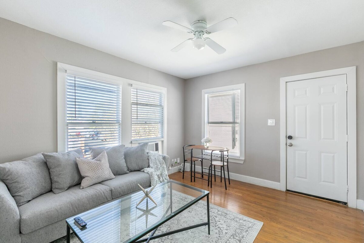 A bright living area features a comfortable gray sofa paired with a glass coffee table on a light rug. Natural light filters through two windows adorned with blinds. A small dining table for two sits near the door, enhancing the inviting atmosphere.