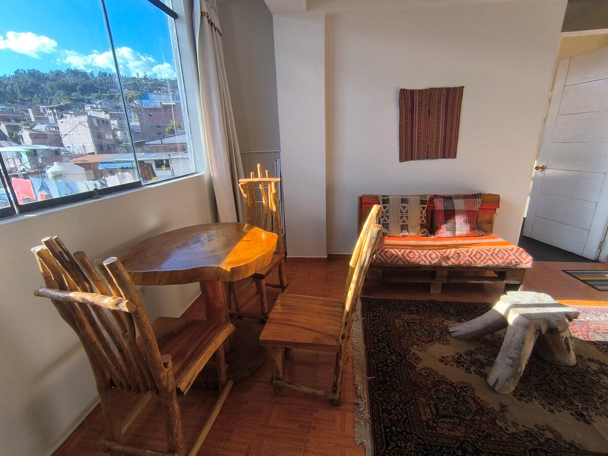 A sunlit corner of the room features a wooden table accompanied by two rustic chairs. A colorful textile artwork adorns the wall. A low seating area with vibrant cushions is positioned beside a patterned rug, creating an inviting space for relaxation.