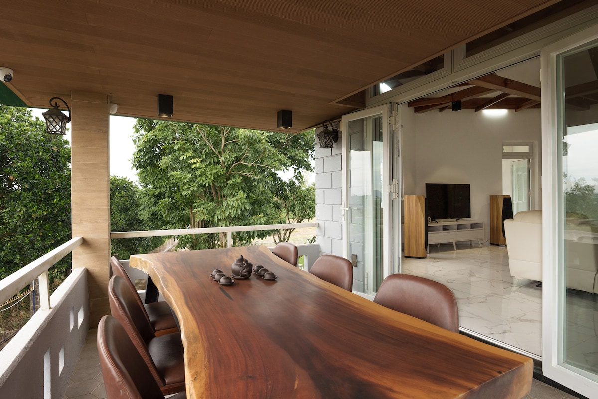 An outdoor dining area features a long wooden table with dark, comfortable chairs, perfect for gatherings. Large sliding glass doors open to a bright living space, while lush greenery is visible in the background, adding to the serene environment.