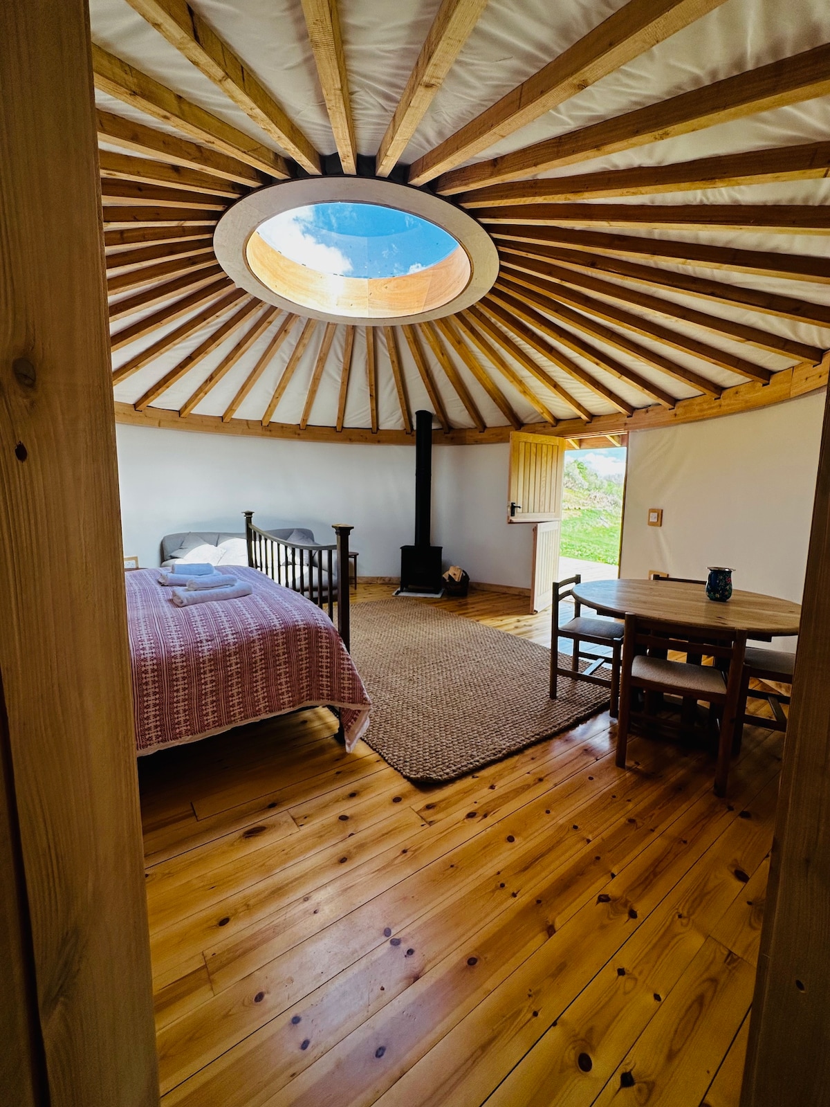A roundhouse cabin showcases a spacious sleeping area with a super-king bed, cozy bedding, and large skylight above. A wood burner is positioned nearby, accompanied by a small dining table and chairs. Natural light highlights polished wooden floors, enhancing the serene interior atmosphere.