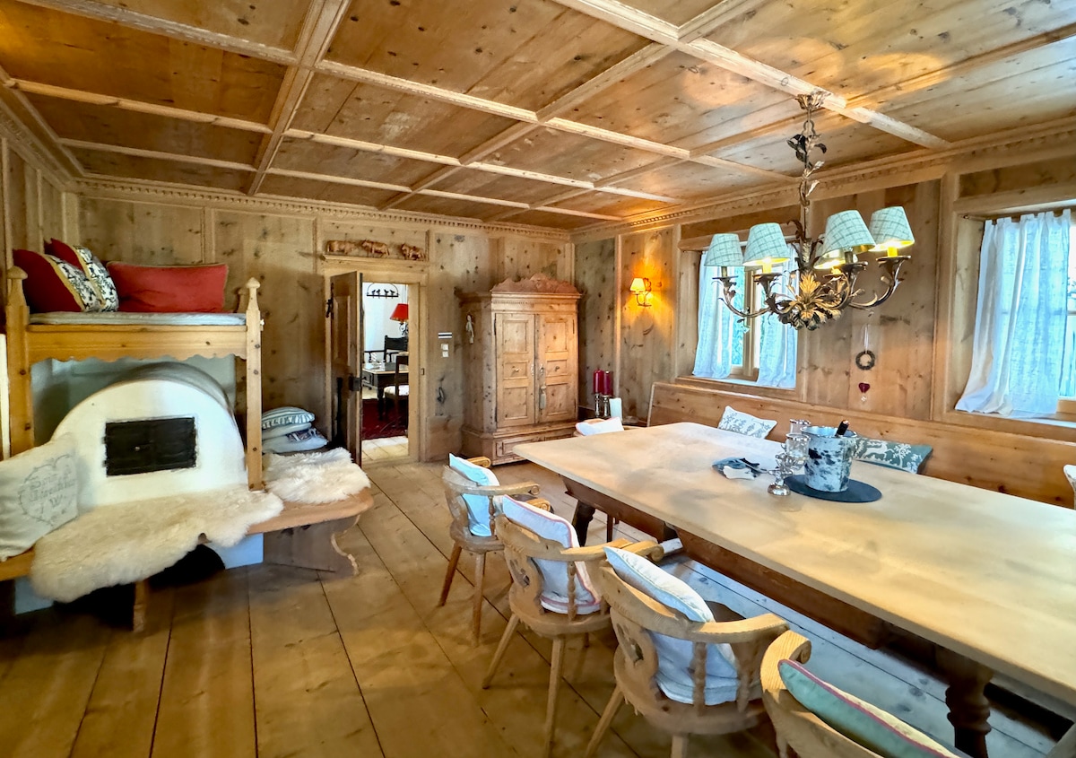 A warm and inviting dining area showcases a large wooden table surrounded by several chairs. Cozy elements include a traditional fireplace with a wooden bench and decorative lighting from a chandelier. Soft textiles and natural light from window treatments complement the rustic wooden walls.
