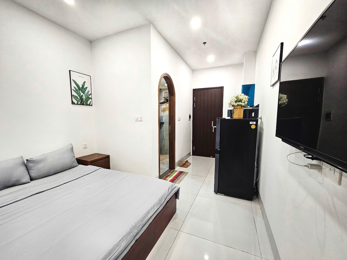 A simple and modern room features a spacious bed with light gray bedding, complemented by a wooden nightstand. A flat-screen television is mounted on the wall opposite the bed. An arched doorway leads to an entry area, and a small refrigerator is present along the wall.