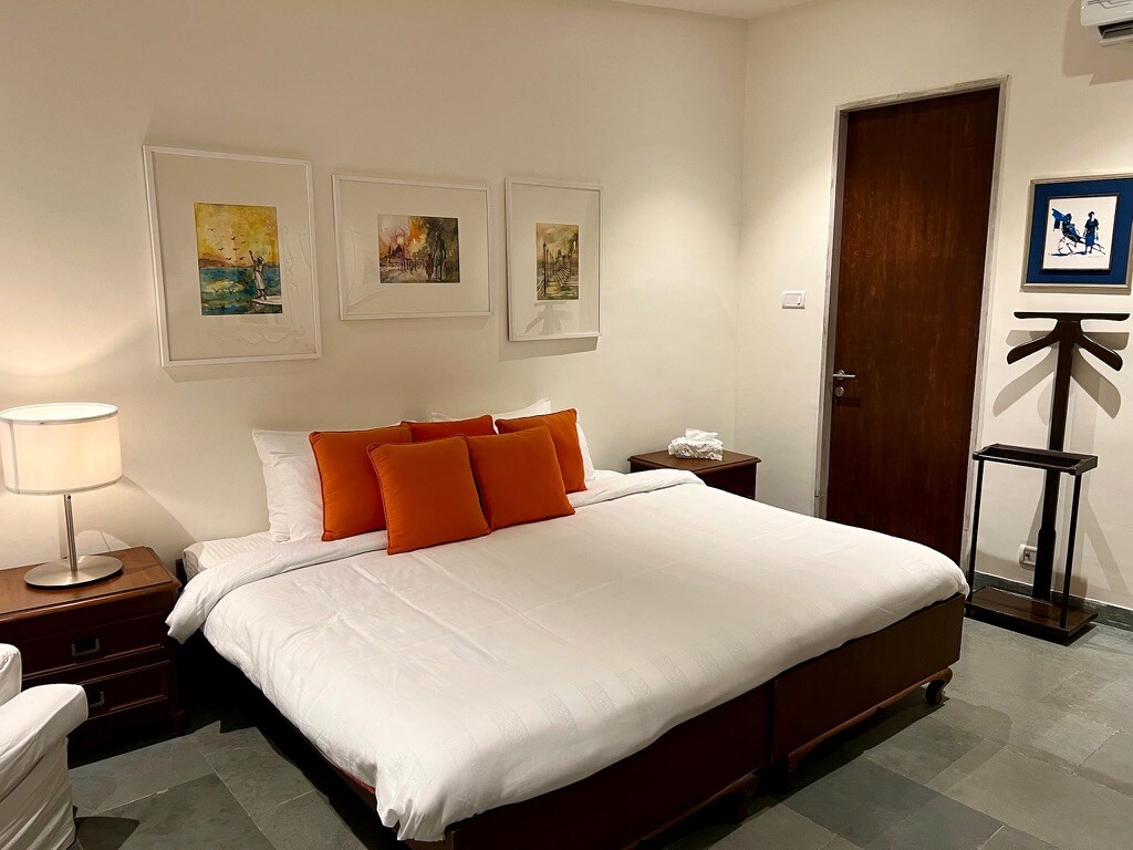 A spacious room features a deluxe king bed with white linens and orange accent pillows. Framed art decorates the walls, and a bedside lamp provides soft illumination. A wooden door and a small side table with an additional item can be seen in the background.