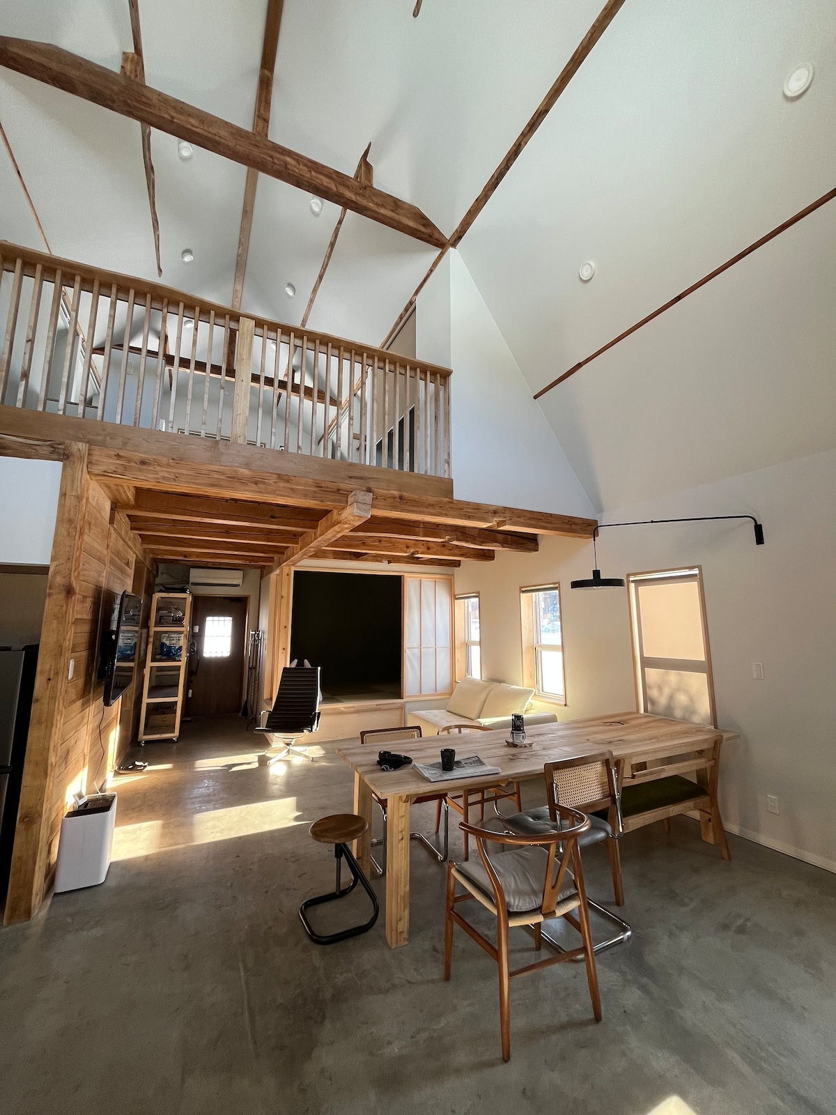 A spacious interior showcases high ceilings with exposed wooden beams, contributing to an airy atmosphere. A large wooden dining table is centrally positioned, surrounded by simple stools. Natural light enters through windows, illuminating the area and highlighting the minimalist yet warm design.