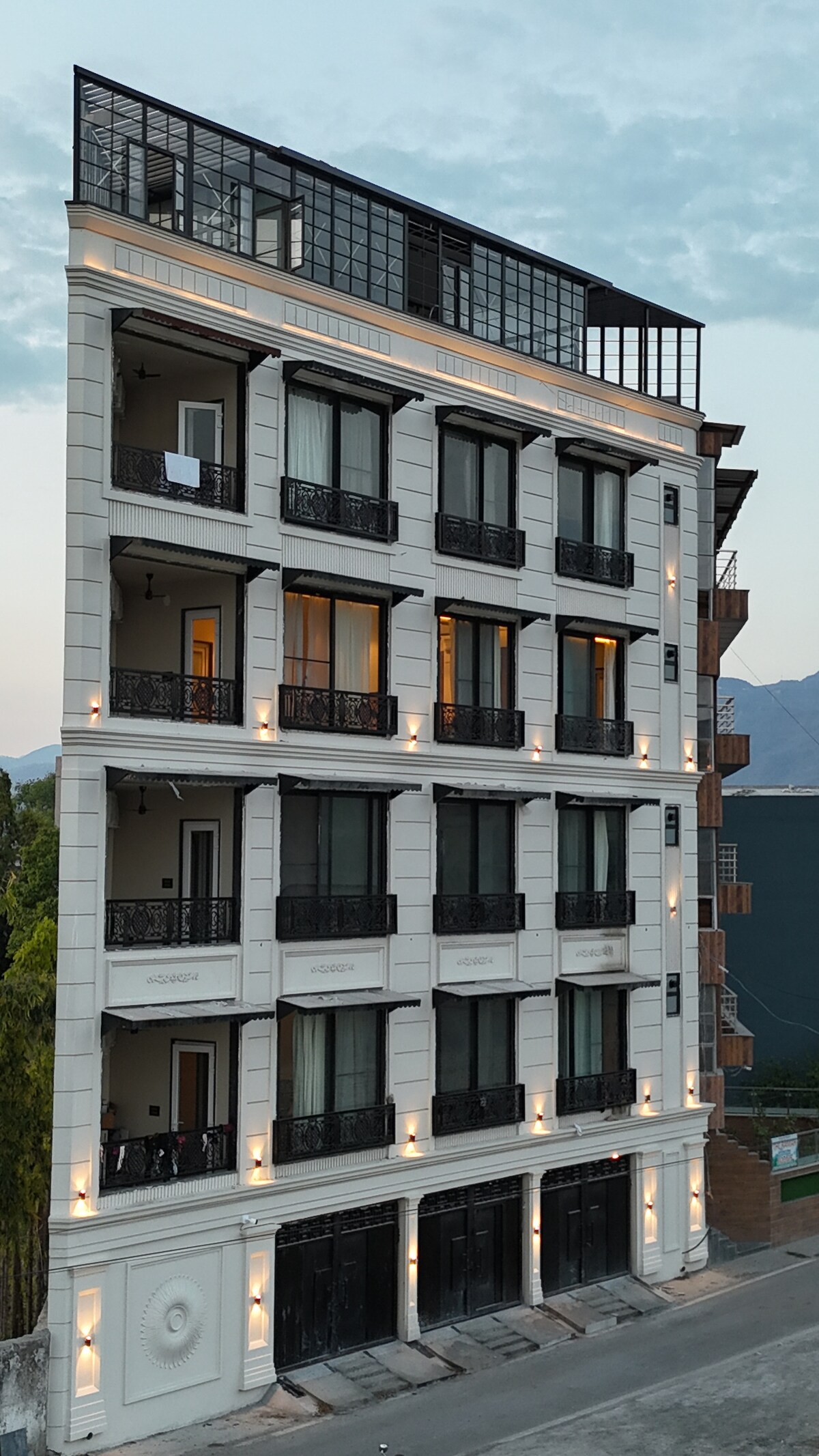 The elegant five-story building features a classic façade with large windows and balconies, enhancing its architectural appeal. Soft lighting highlights the structure's contours as it stands against a backdrop of mountains and a dusky sky.