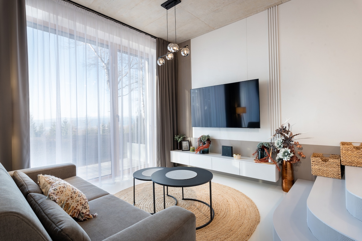 A modern living area features a comfortable sofa facing a wall-mounted smart TV. Large windows adorned with sheer curtains allow natural light to fill the space. A round coffee table is centered on a textured area rug, enhancing the room's inviting design.