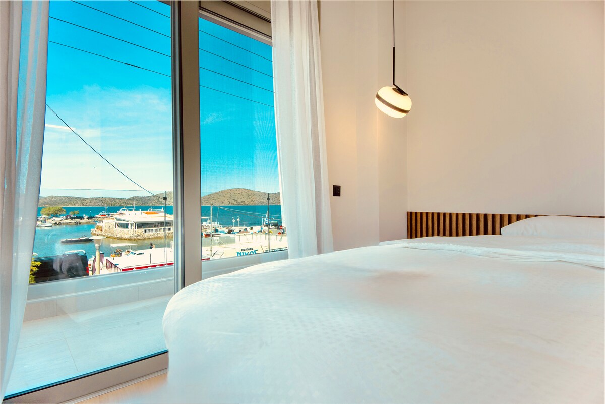 A comfortable bedroom is presented with a spacious bed featuring crisp white linens. Large glass doors open to a stunning view of the marina, where boats are docked against a backdrop of the tranquil water and distant hills. Soft natural light fills the room.