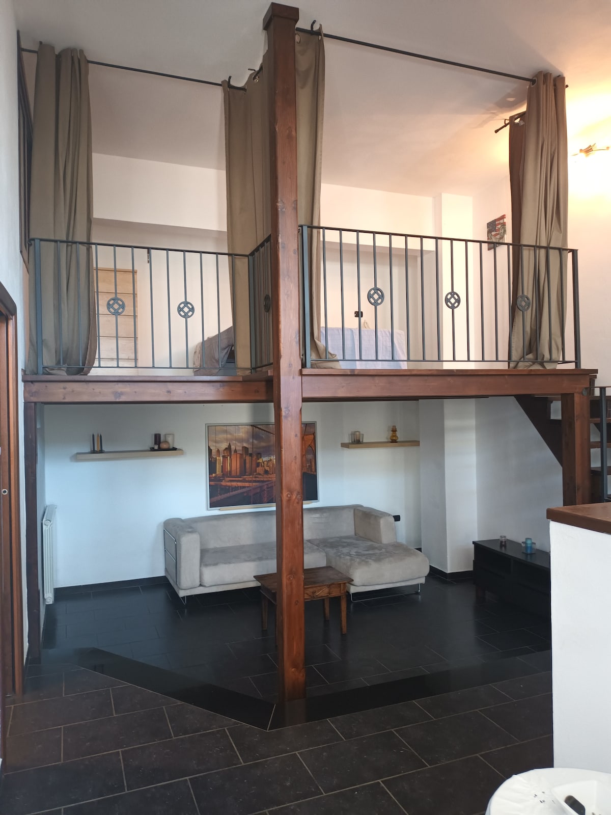 The loft features a spacious open area with high ceilings, showcasing exposed wooden beams. A comfortable sofa is positioned below a sleeping loft, which includes a cozy sleeping area surrounded by soft drapes. Decorative shelves and a small table add functional details to the living space.