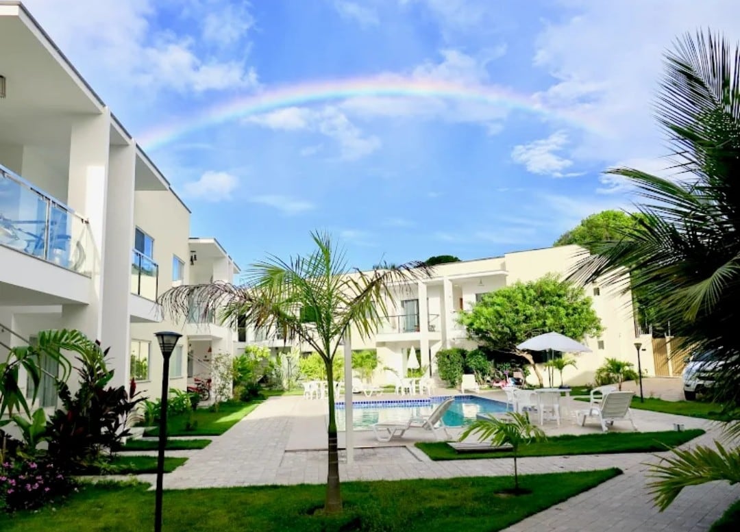 A serene courtyard is visible, featuring a well-maintained pool surrounded by lounge chairs. Lush greenery and tropical plants frame the area. A rainbow arches over the sky, providing a colorful backdrop to the modern buildings that house the accommodations.