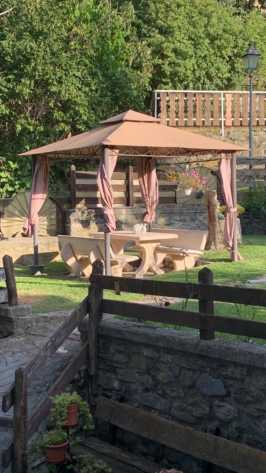 A shaded gazebo is set on green grass, featuring a round table surrounded by wooden chairs. Delicate drapes add softness to the structure, while nearby plants provide a natural touch. Stone walls and a rustic fence frame the charming outdoor area.