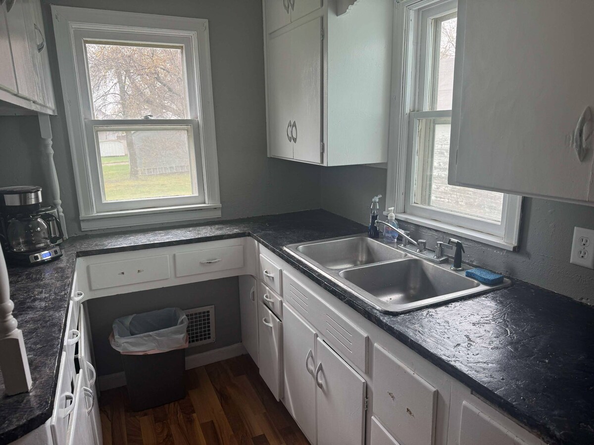 A functional kitchen space is presented with white cabinetry and a dark countertop. Sunlight filters through two windows, highlighting the double sink and various appliances, including a coffee maker. The floor is finished in warm wood tones, contributing to an inviting environment.
