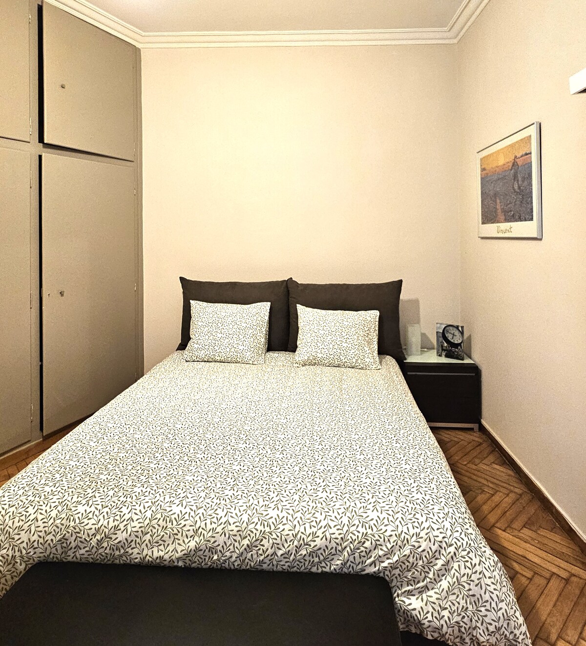 A cozy bedroom is furnished with a spacious bed dressed in light-coloured bedding featuring a subtle pattern. Two decorative pillows are placed against a neutral wall. A nightstand with a clock is positioned beside the bed, while a closet offers storage space. Natural light fills the room.