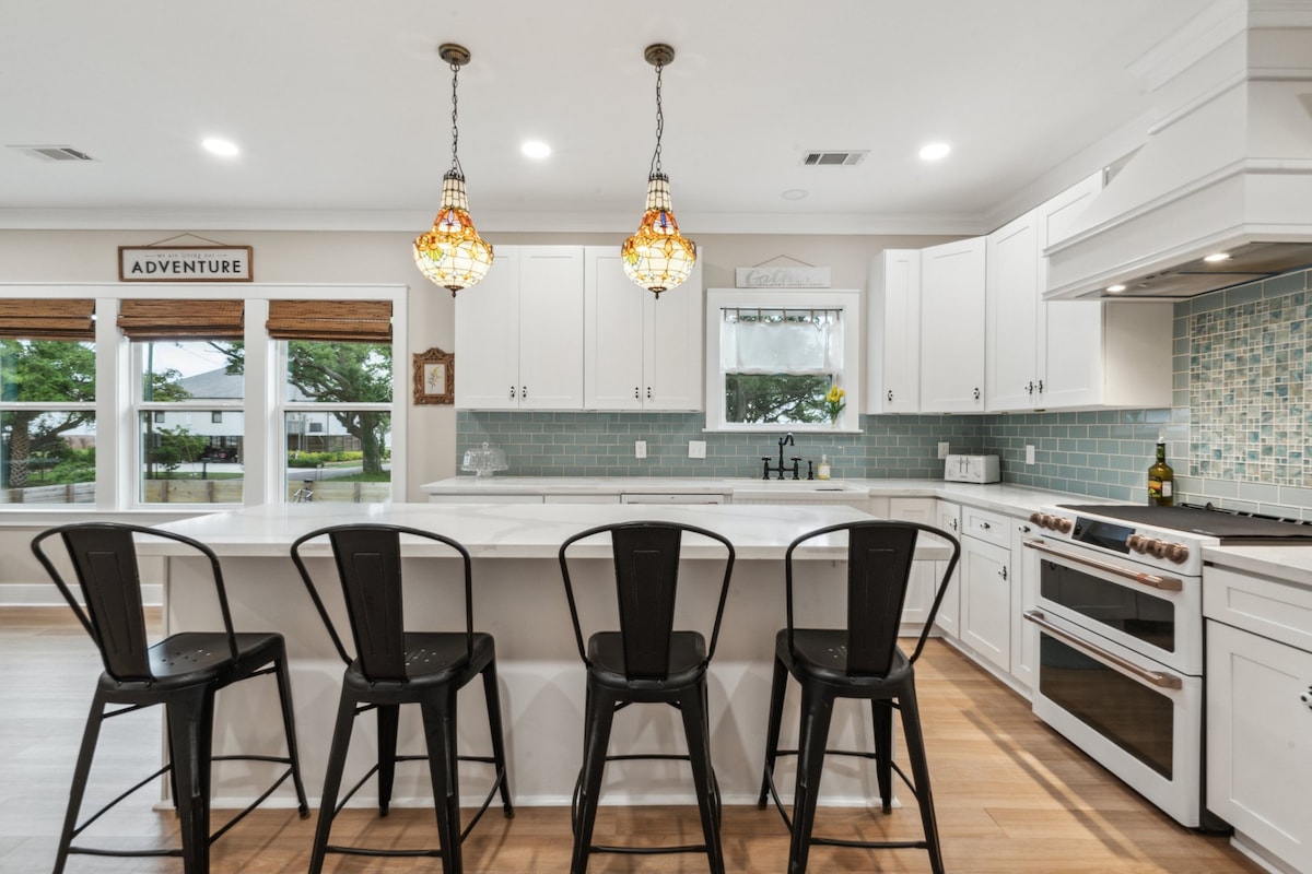 A modern kitchen features a large island with four black chairs arranged around it. Pendant lights hang above, providing soft illumination. The backsplash is adorned with blue tiles, while large windows offer natural light and views of the outdoor surroundings.